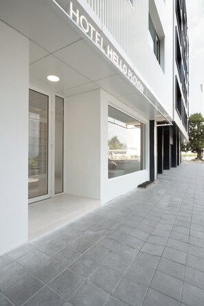 Une entrée d’hôtel moderne avec une façade en verre transparent, une enseigne indiquant « HÔTEL HELLO PLOVIDY » et un trottoir dallé menant à l’entrée.