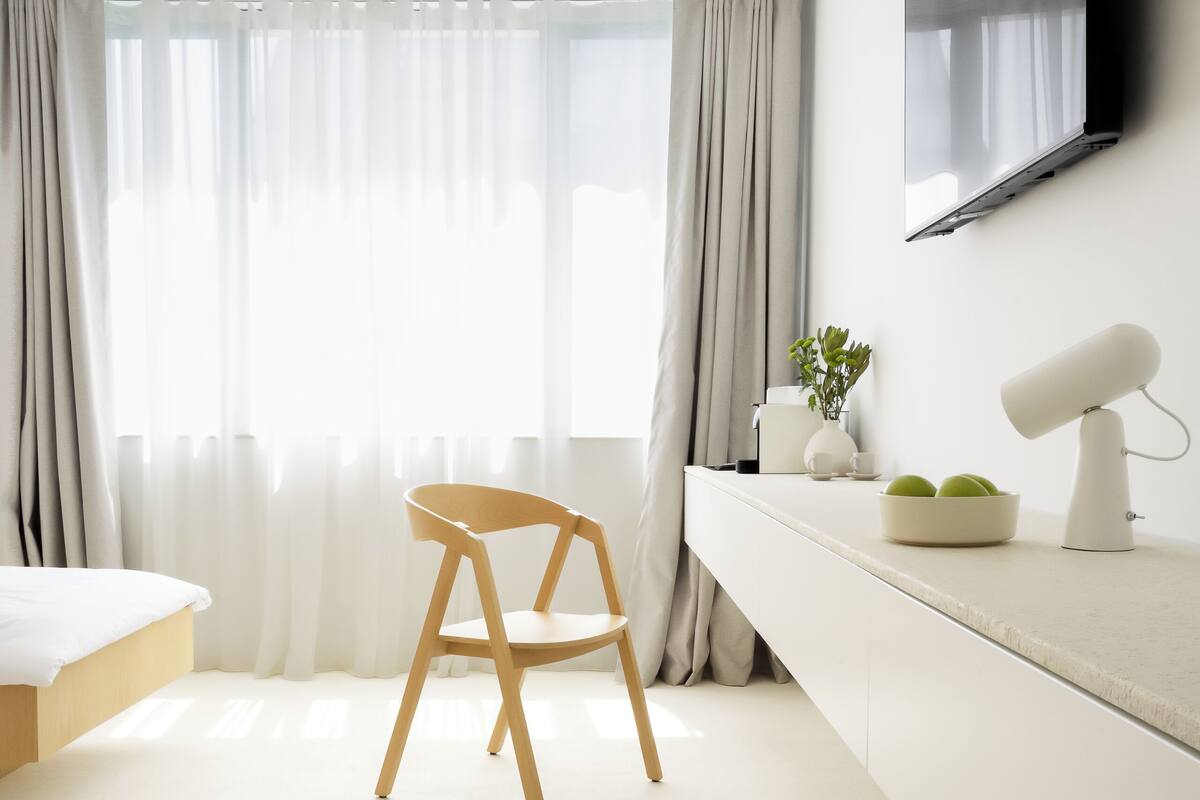 A minimalist hotel room with a bed, a desk with a lamp, a chair, and a TV mounted on the wall.