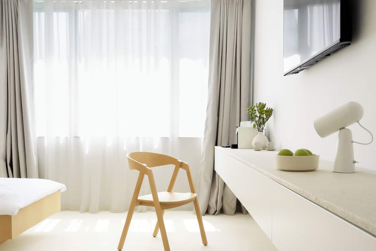 A minimalist hotel room with a bed, a desk with a lamp, a chair, and a TV mounted on the wall.