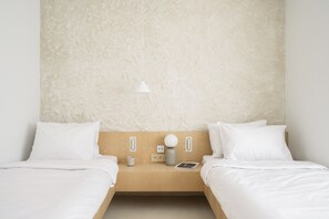 A minimalist hotel room with two single beds, a wooden headboard, a bedside table with a lamp, a cup, and a book. - Hotel Hello Plovdiv (Plovdiv)