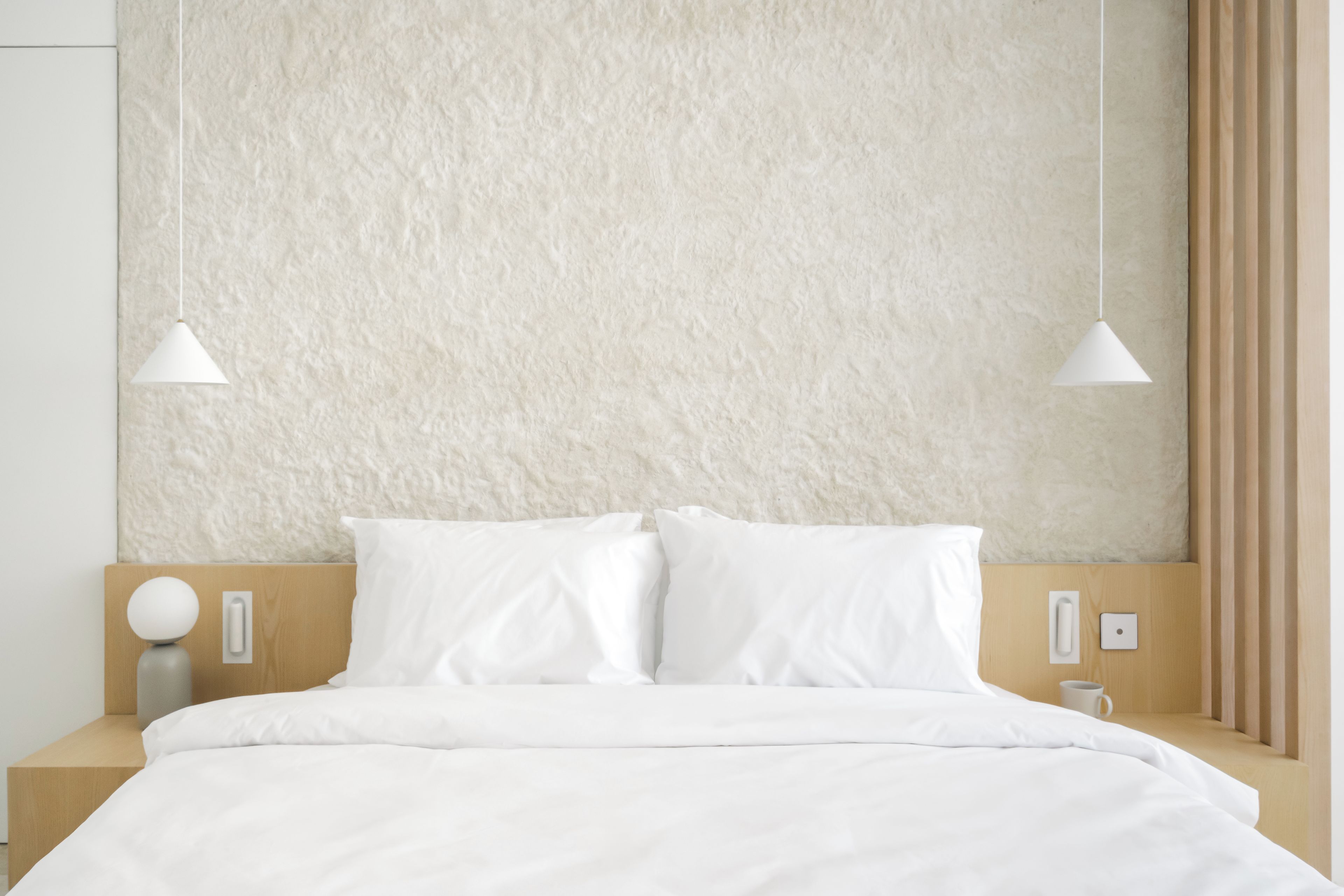 A modern hotel room with a textured beige wall, a wooden headboard, white bedding, and two pendant lights.