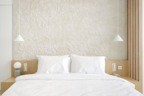 Une chambre d’hôtel moderne avec un mur beige à texture, une tête de lit en bois, du linge de lit blanc et deux lampes suspendues.