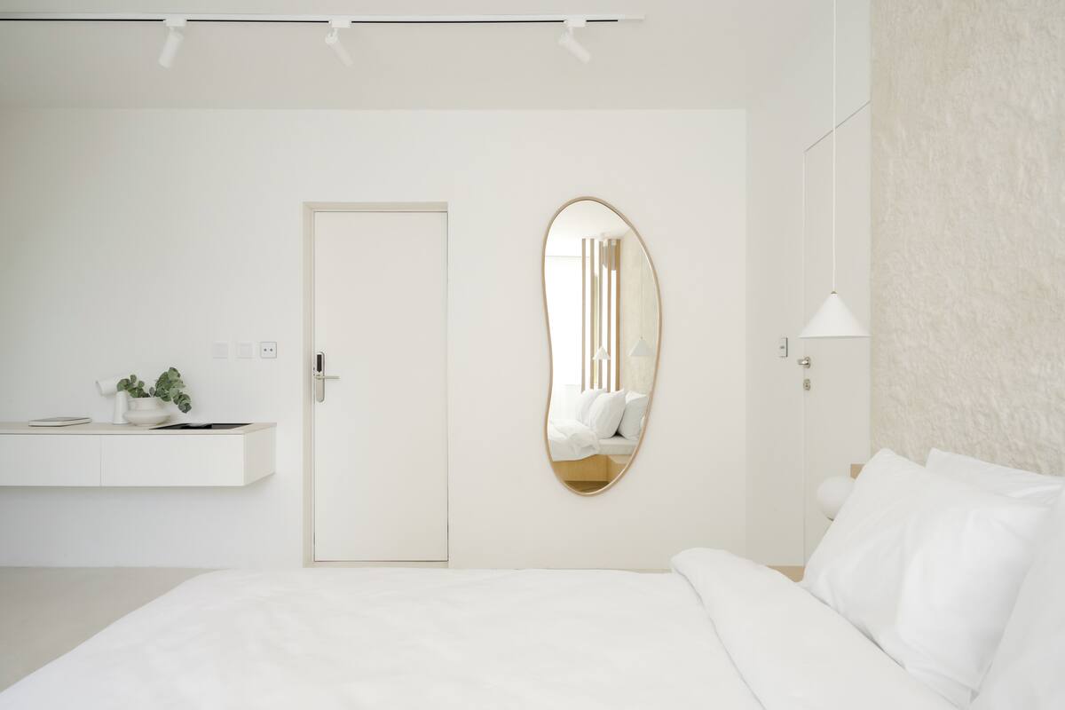 A minimalist bedroom with a large bed, a bedside table with a plant, a door, and a wall-mounted oval mirror.
