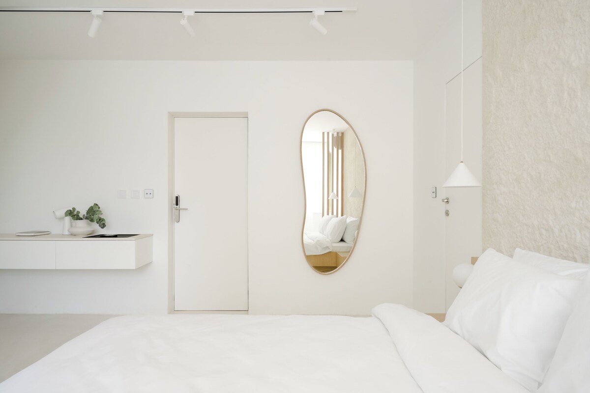 Une chambre à coucher au style minimaliste, avec un grand lit, une table de chevet agrémentée d’une plante, une porte et un miroir ovale fixé au mur.