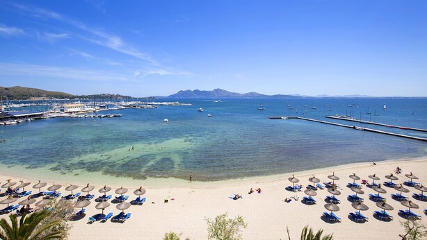 Ein Strand mit klarem Wasser, einer Marina und einem sandigen Ufer mit Sonnenschirmen und Liegestühlen.