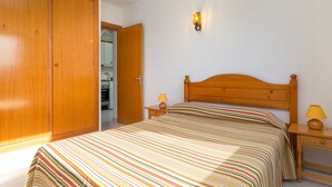 Un dormitorio con un armario de madera, una cama con colcha a rayas, dos mesitas de noche con lámparas y un aplique en la pared.