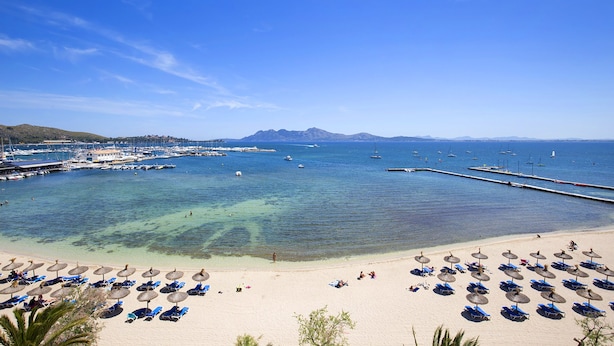 Una playa con aguas cristalinas, un puerto deportivo y una orilla arenosa con sombrillas y sillas de playa.