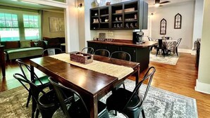 A dining area with a wooden table and chairs, a kitchen with dark cabinets and hanging lights, and a seating area with a green sofa and windows.