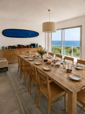 A dining area with a long wooden table, chairs, and a view of the ocean through large windows.