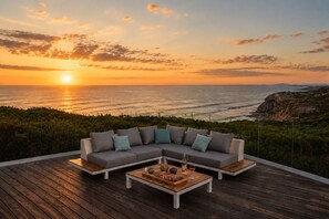 A modern outdoor seating area with a wooden deck, a gray sectional sofa with pillows, and a small table with a bottle and glasses.