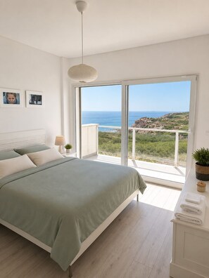A bedroom with a bed, a bedside lamp, a small plant, and a view of the sea.