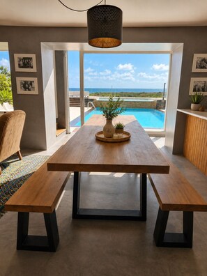 A modern dining area with a wooden table and benches, a hanging light fixture, and a view of a swimming pool and ocean. - Modern Eco-Responsible House with Ocean View, Swimming Pool, Surfing, Golf, Tennis (Lourinha)