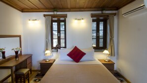 A hotel room with a wooden ceiling, two windows with curtains, a bed with a red pillow, bedside lamps, a desk with a chair, and a small plant on the desk.