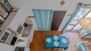 Cocina compacta con comedor, que incluye una mesa azul para cuatro personas, un refrigerador pequeño y una ventana con cortinas.