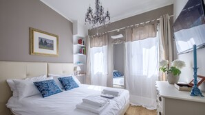 Ein ordentlich bezogenes Bett mit weißen Leinen und blauen Kissen, eine Kristallleuchter, ein Bücherregal mit Büchern und ein Fenster mit durchsichtigen Vorhängen.
