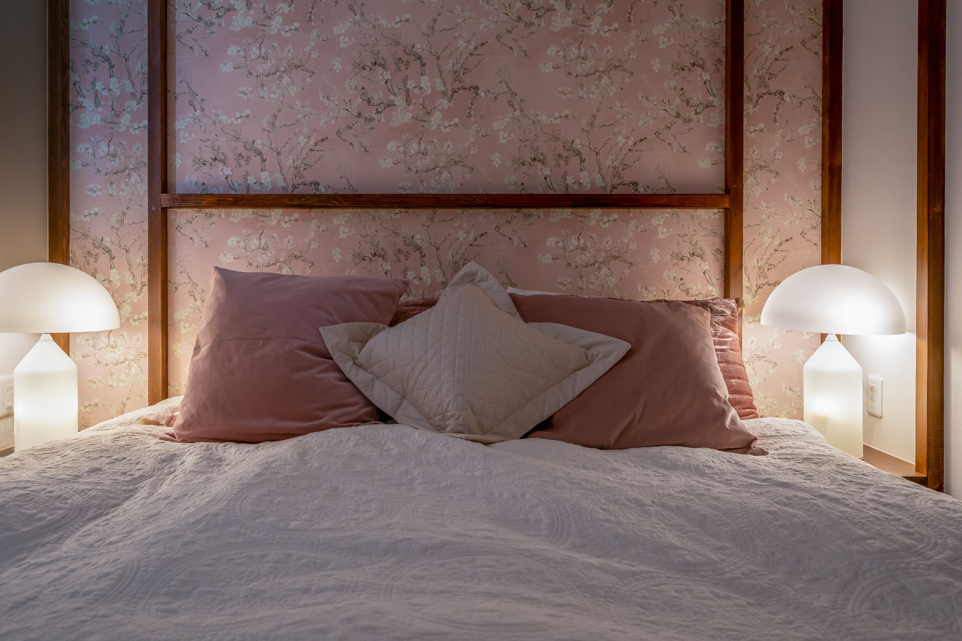 A neatly made bed with a quilted white duvet, pink and white pillows, and a wooden headboard with floral wallpaper.