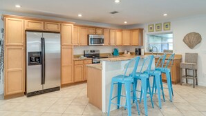 Una cocina moderna con armarios de madera, un refrigerador de acero inoxidable, un microondas y una cafetera. Hay tres taburetes azules en la isla.