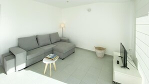 Una sala moderna con un sofá seccional gris, una pequeña mesa blanca con libros, una lámpara de pie, un televisor sobre un mueble blanco y una cesta de mimbre.