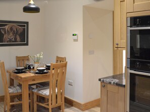 Un coin repas avec une table en bois dressée pour quatre personnes, un tableau accroché au mur et une cuisine équipée d’un four encastré.