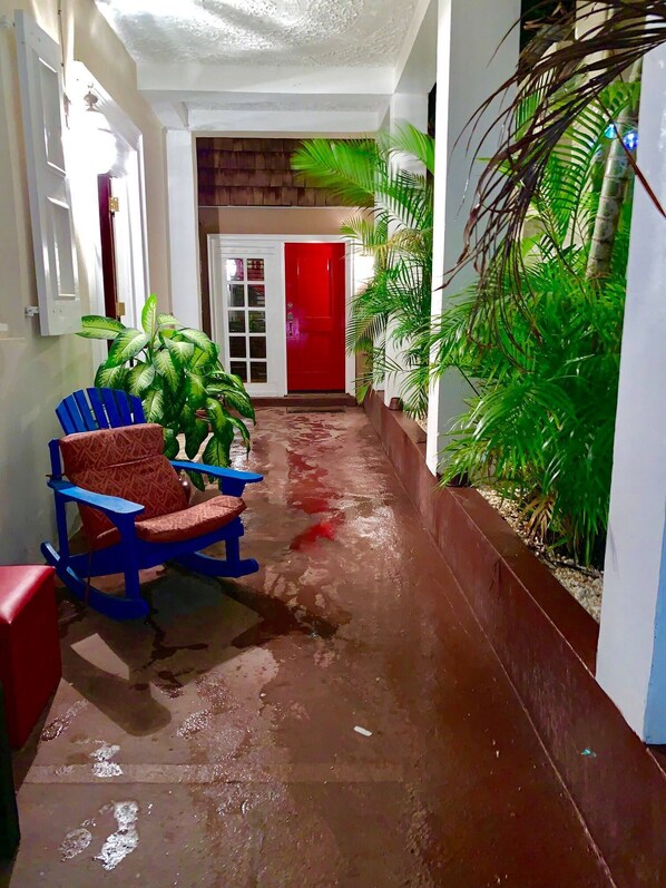 Un couloir étroit avec une porte rouge, une chaise bleue et des plantes en pot.