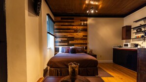 Une chambre à coucher avec une tête de lit en bois, un lit, une table de chevet avec un vase, et un coin cuisine équipé d’un four à micro-ondes et de placards.