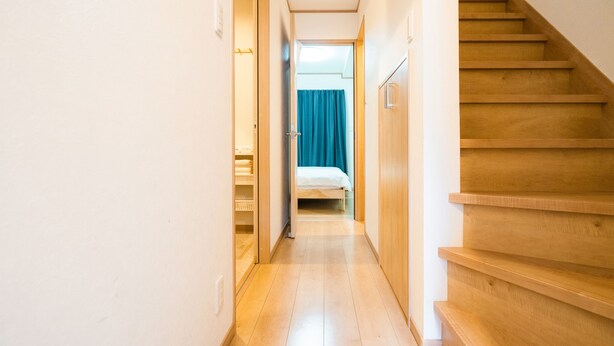 Um corredor estreito com escada de madeira à direita, que leva a um quarto com cama e cortina azul.