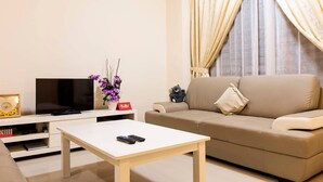 Een woonkamer met een beige bank, een witte salontafel, een flatscreentelevisie en een kleine sierplant.