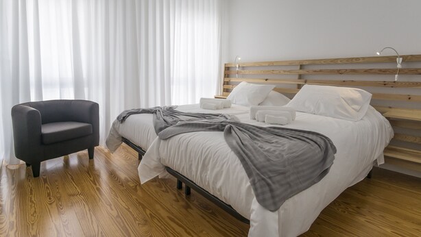 Ein modernes Schlafzimmer mit einem Holz-Kopfteil, einem Bett mit weißer Bettwäsche, einer grauen Decke und einem schwarzen Sessel.