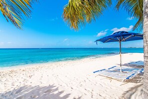 A beach with white sand, clear blue water, and a blue umbrella providing shade.