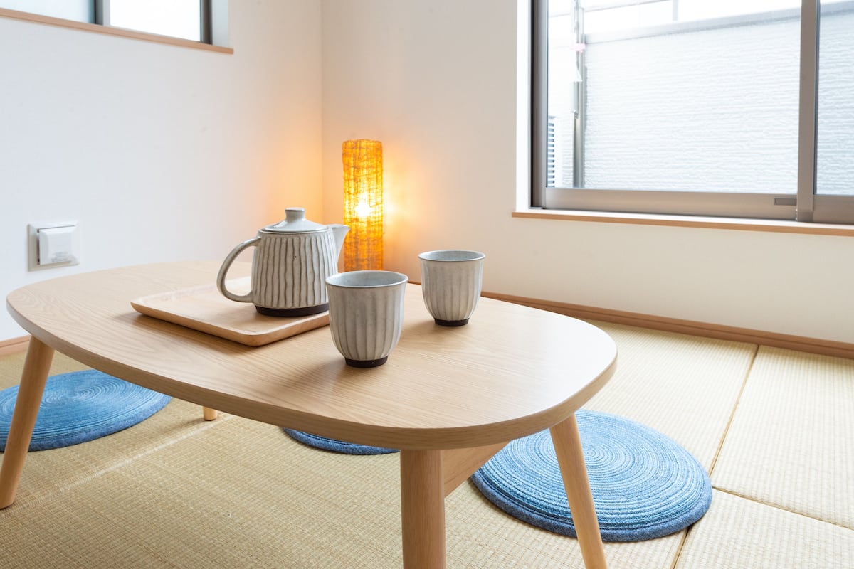 Une table ronde en bois, sur laquelle se trouvent un plateau et deux tasses en céramique, une grande lampe au design original et un tapis bleu tissé.