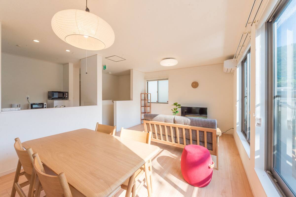 Un salon moderne avec une table à manger en bois, un pouf rouge, un canapé gris et une grande fenêtre laissant entrer la lumière naturelle.