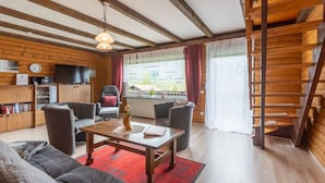 Una habitación acogedora de estilo cabaña, con techo de madera, zona de comedor con mesa y sillas, sofá, televisor y estantería.