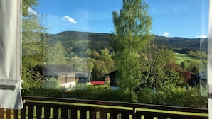 Vista desde una ventana con baranda de madera, que se extiende sobre un paisaje rural con árboles, una colina y algunos edificios.
