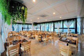 A spacious dining area with multiple tables and chairs, large windows with curtains, and hanging greenery. - Angel Hotel Nha Trang (Nha Trang)