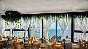 A restaurant with tables and chairs arranged for dining, large windows with white curtains, and a view of the ocean. - Angel Hotel Nha Trang (Nha Trang)