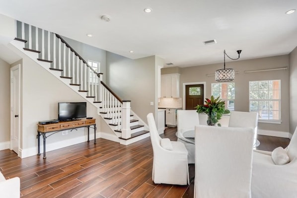 A spacious living area with a wooden floor, a staircase, a dining table with chairs, and a television on a stand.