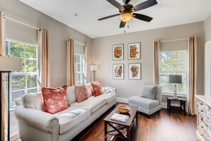 A living room with a sofa, armchair, and coffee table, a ceiling fan, and artwork on the walls. - Buttercup - 3BR Home Close to the Convention Center + Walk to Downtown (West Palm Beach)