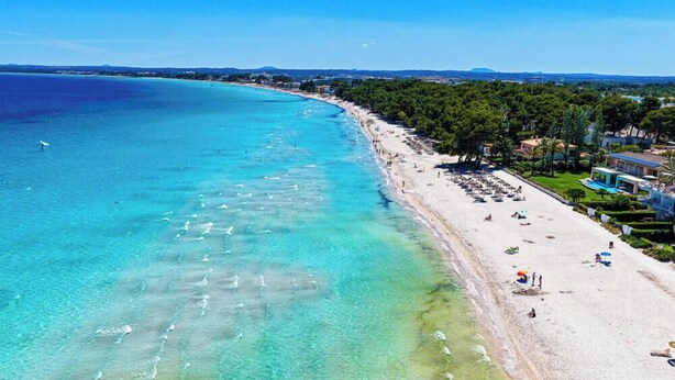 Ein Küstenstrand mit klarem türkisfarbenem Wasser, einem sandigen Ufer und einer Reihe von Gebäuden mit Poolbereich.