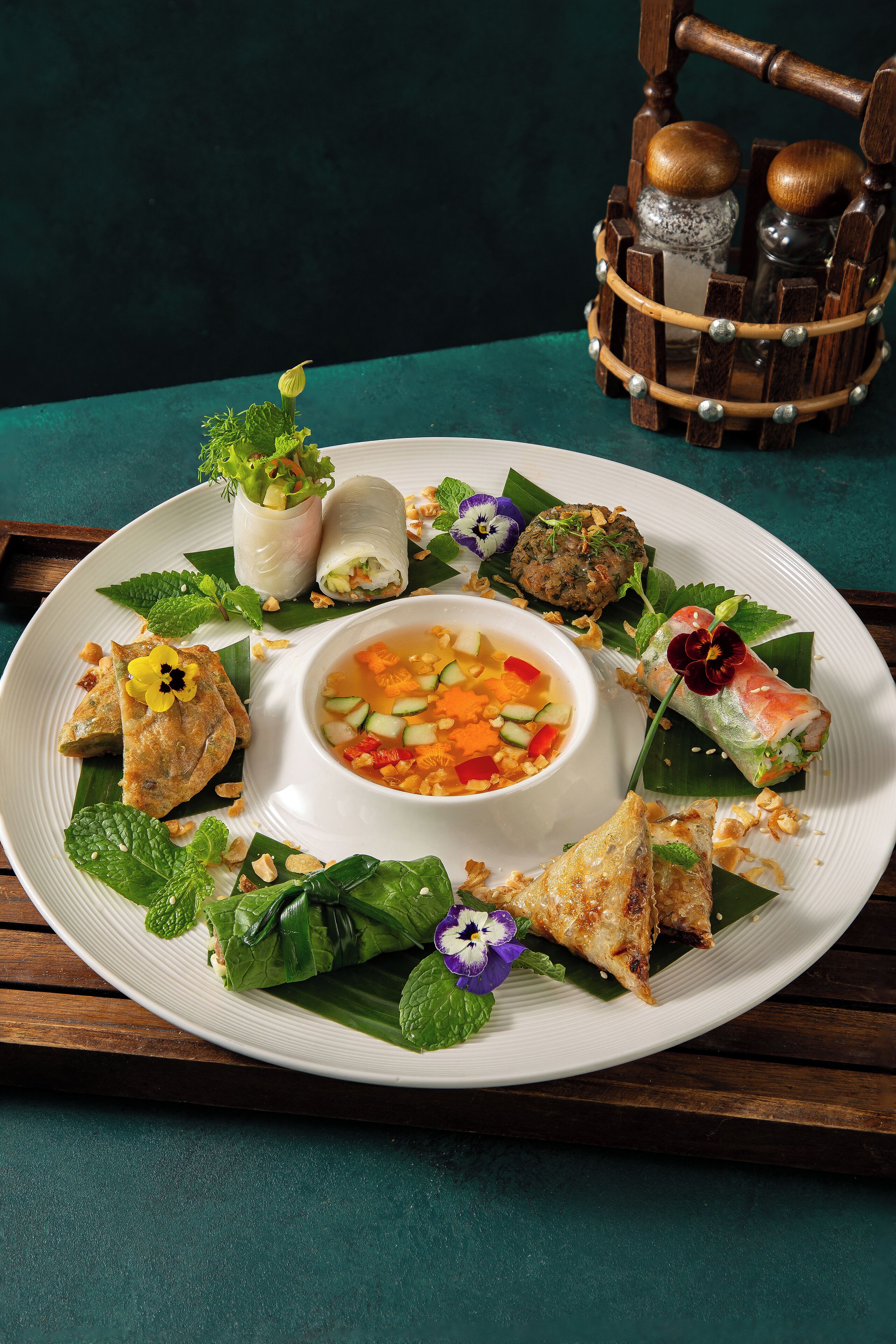 A plate of Vietnamese cuisine with various dishes, including spring rolls, a bowl of soup, and a decorative wooden basket in the background.