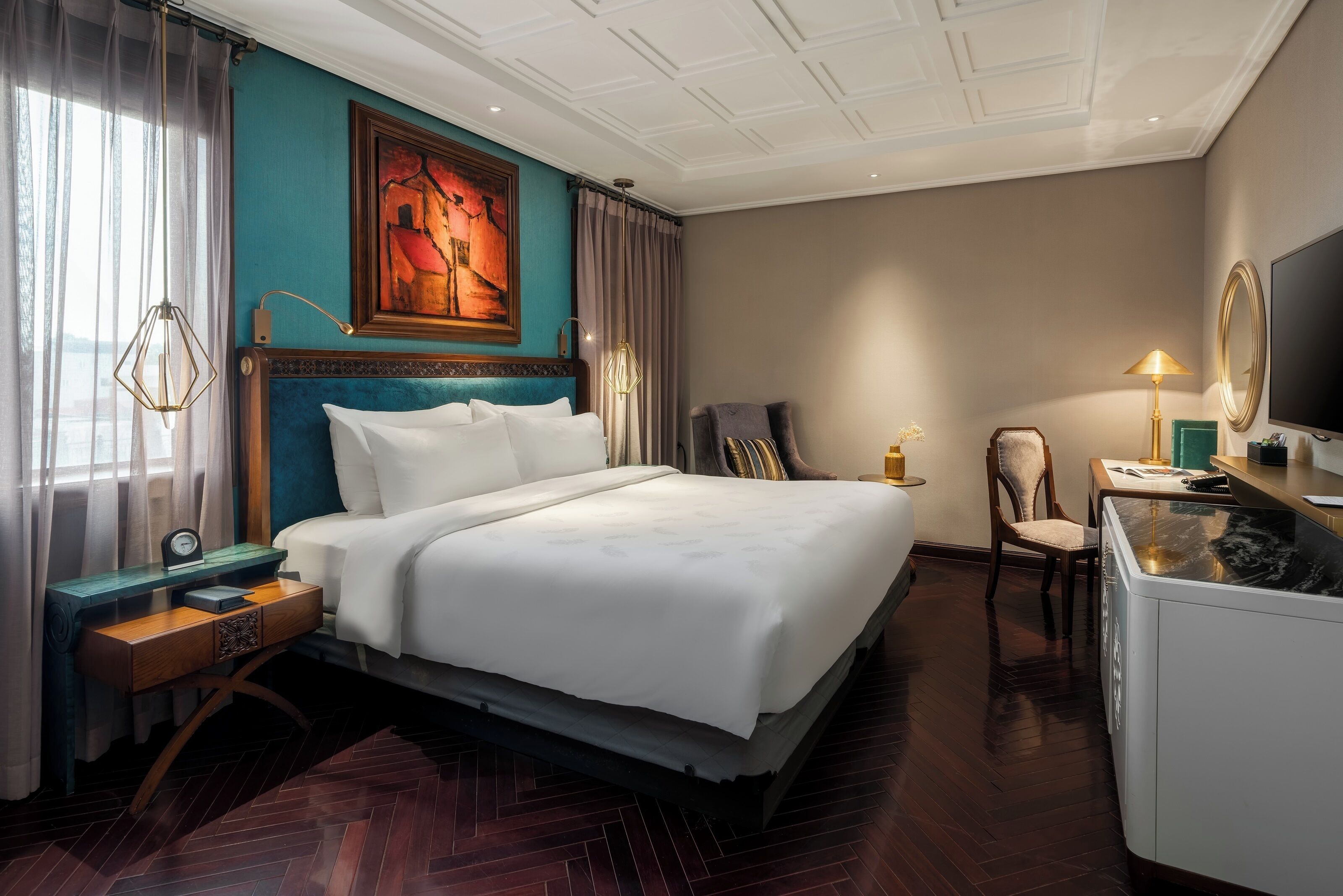 A hotel room with a large bed, a desk with a lamp, a chair, a television, and a painting on the wall.