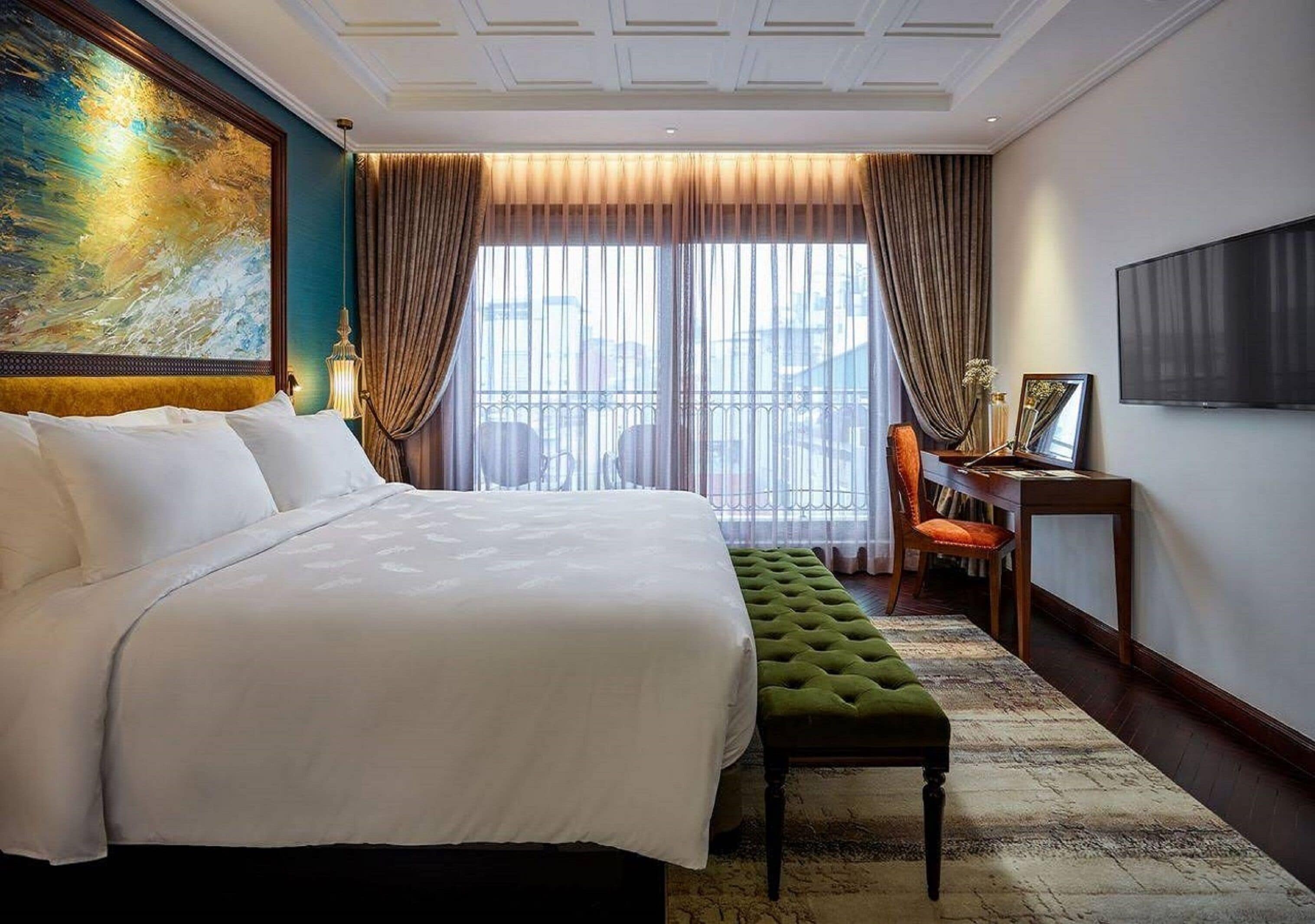 A hotel room with a large bed, a green bench, a desk with a chair, and a large painting on the wall.