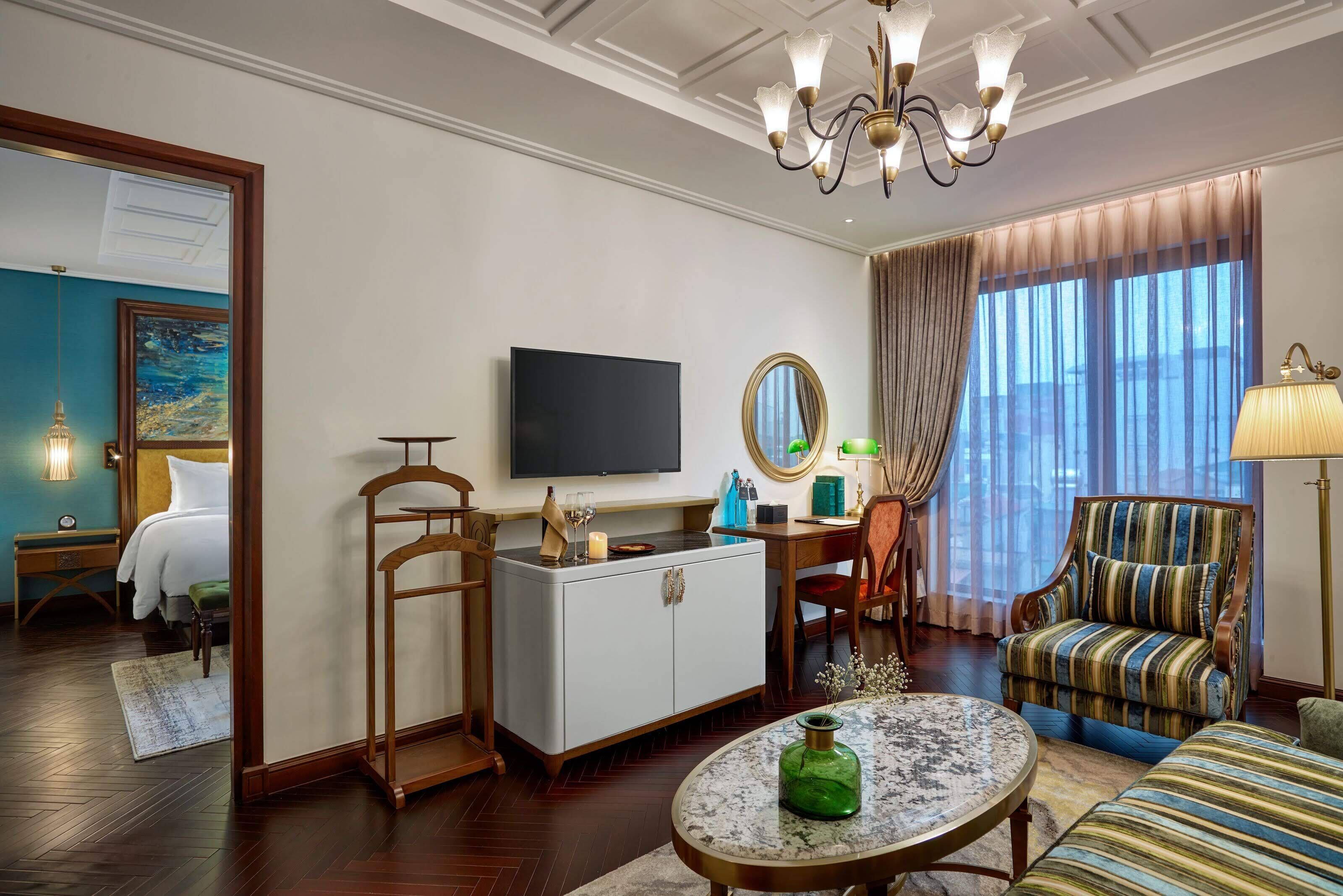 Une chambre d’hôtel comprenant un lit, une télévision, un bureau avec un miroir, un fauteuil à rayures, une table basse ronde et un lustre.