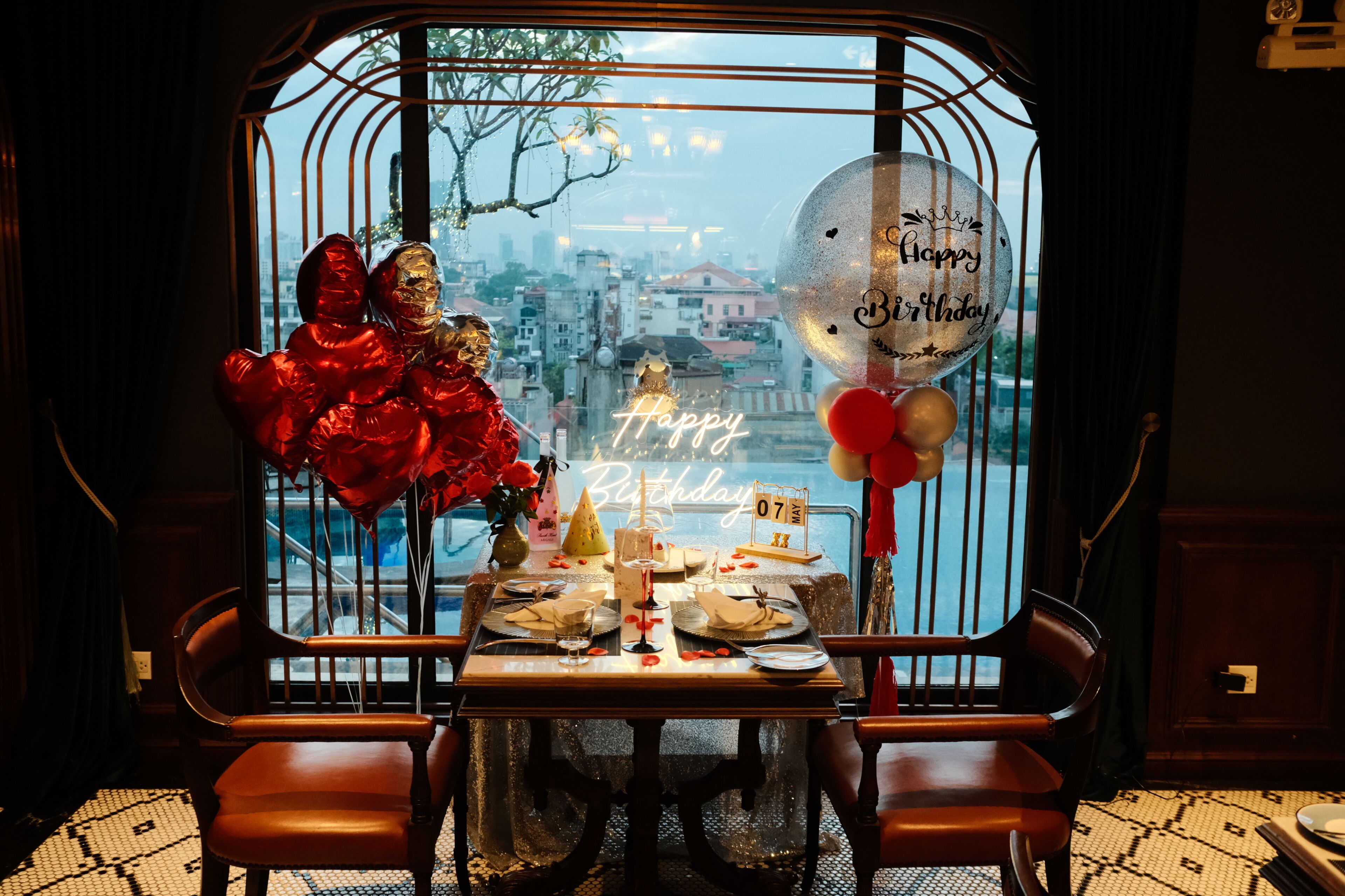 A dining table set for a birthday celebration with a city view, a neon sign, and balloons.