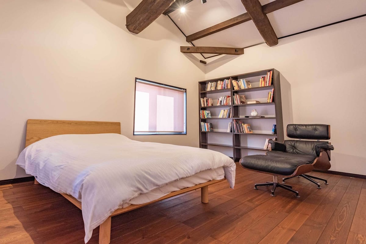 Une chambre à coucher avec un lit en bois, une bibliothèque, un fauteuil en cuir et une fenêtre avec des stores.