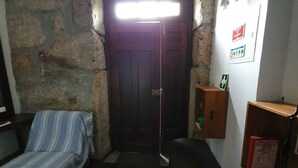Una habitación pequeña e iluminada tenuemente con una pared de piedra, una puerta de madera, una cama con una colcha a rayas, un banco y un estante pequeño con una caja roja.