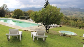 Piscina con dos sillas reclinables blancas y un árbol en un área ajardinada con vistas a montañas.
