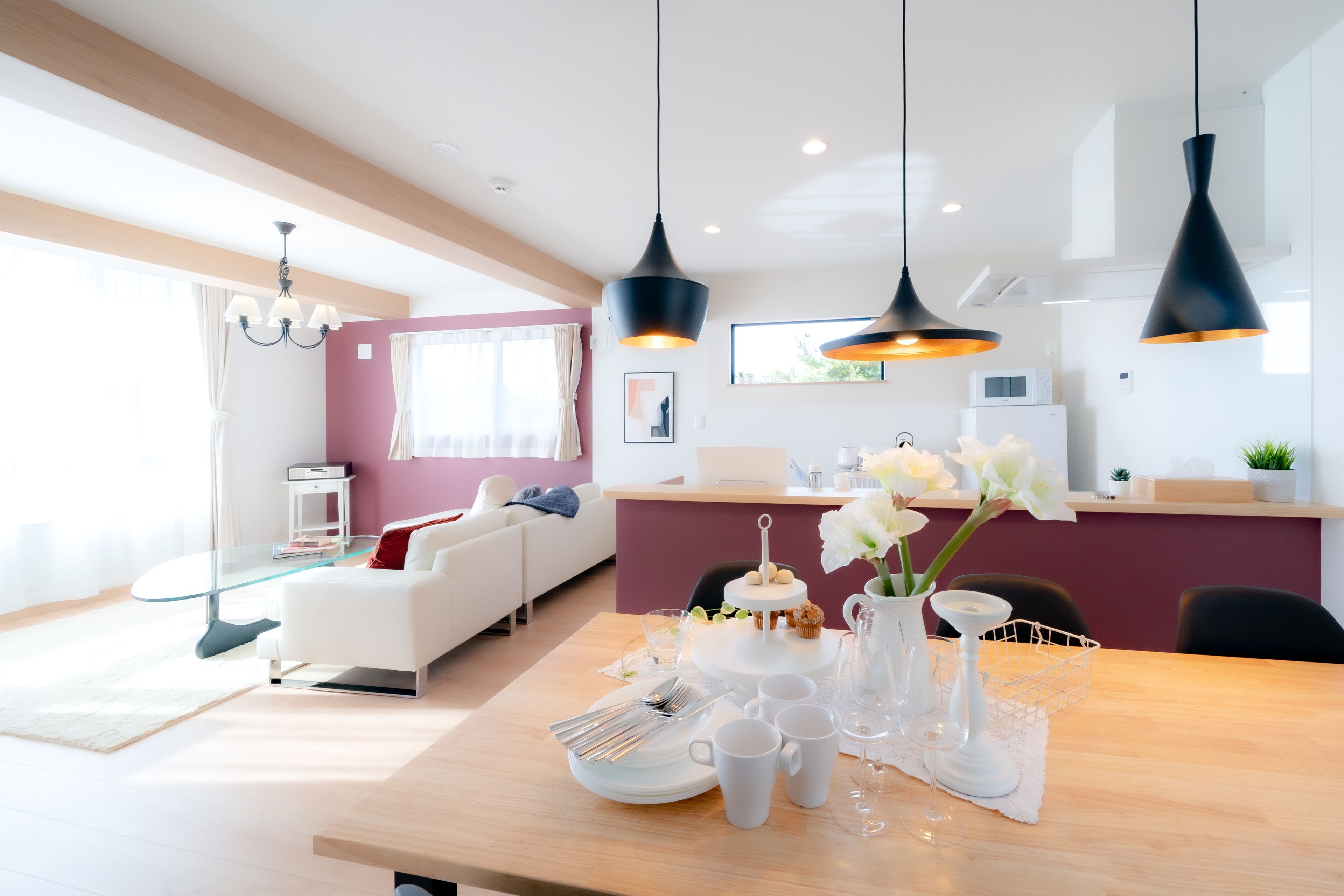 A modern living room with a dining area, featuring a wooden dining table set with white dishes, a vase with flowers, and black pendant lights.