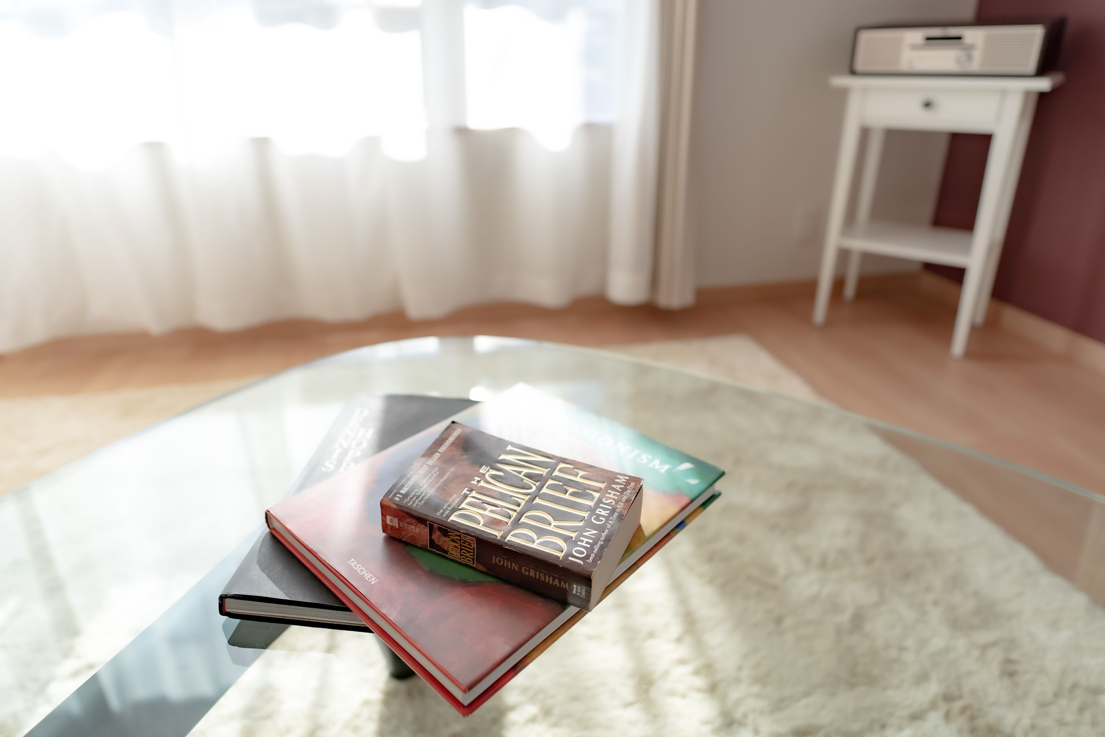 A glass table with books, a white side table, and a window with curtains.