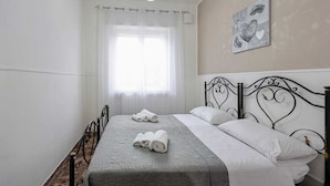Una cama bien hecha con sábanas blancas y toallas enrolladas, un cabecero de metal negro y un ventanal con cortinas translúcidas.