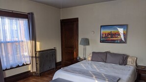 Ein Schlafzimmer mit einem Bett, einem Nachttisch, einer Lampe, einer Heizung, einem Fenster mit Vorhängen, einem Bild an der Wand und einer Bank.